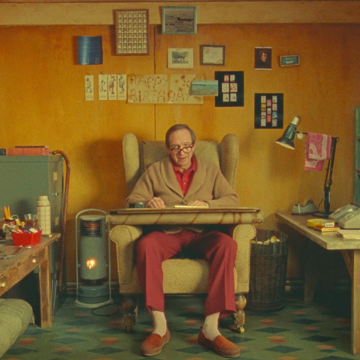 an older man sits in an armchair in a cozy living room, reading a book. he is wearing red pants and a brown sweater, and the room is decorated with vintage furniture and artwork. there is a fireplace in the background, and a lamp on a table beside the man. the floor is tiled with a green and white pattern. the man is smiling and looks relaxed and content.