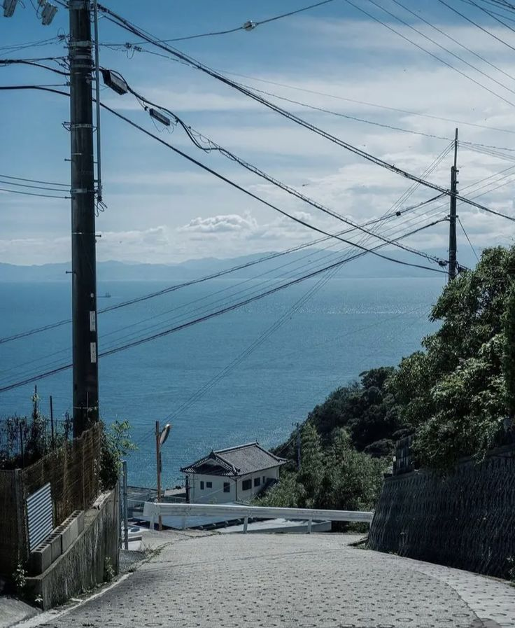 a view from the top of a steep, cobbled street looking down towards a building and the ocean. on the left side of the street is a fence and a concrete retaining wall. on the right side is a stone retaining wall and trees. a guard rail is located in front of the building. telephone poles and power lines cross above the street, obstructing the view of the sky and ocean.