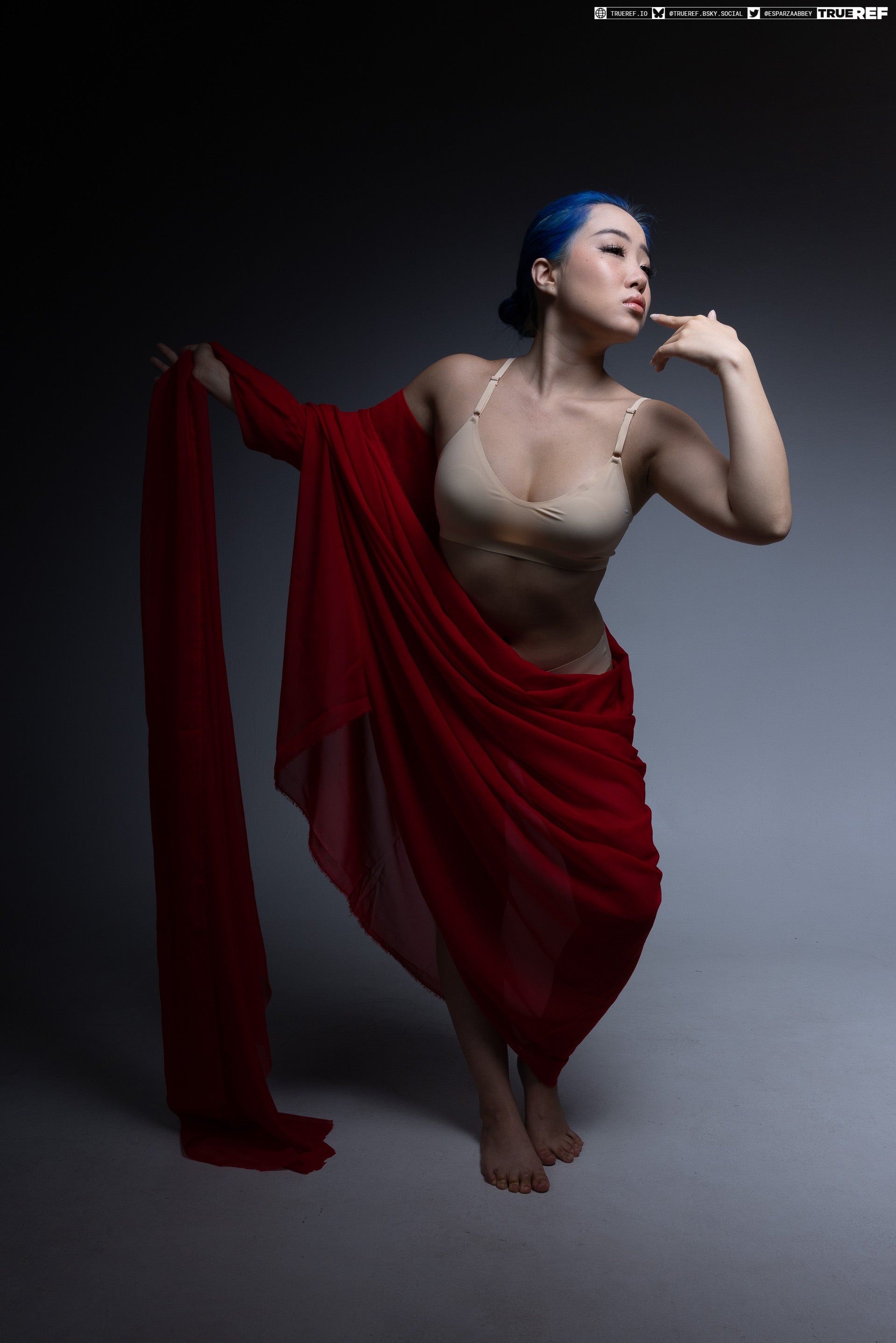 a woman with blue hair is posing in a studio. she is wearing a bra and has a long piece of red fabric draped around her body. she is barefoot and has one hand raised to her face. the background is a dark gray.