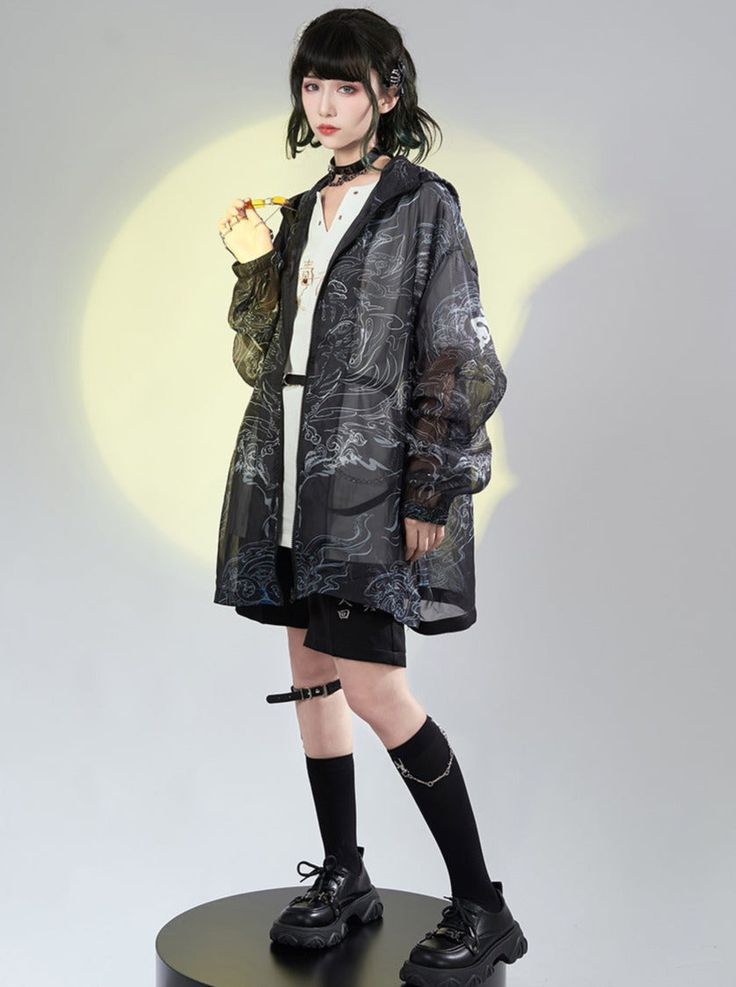 a fullbody studio shot of a young asian woman modeling a black sheer oversized jacket with a wave print. the jacket is styled over a white shirt and black shorts. the model is accessorized with a black choker, silver chain leg jewelry, black socks, and chunky black platform shoes. her hair is styled in a dark bob. the overall style is a mix of japanese streetwear influences and dark, edgy fashion.  the background is a neutral light gray with a subtle yellow highlight behind the model.