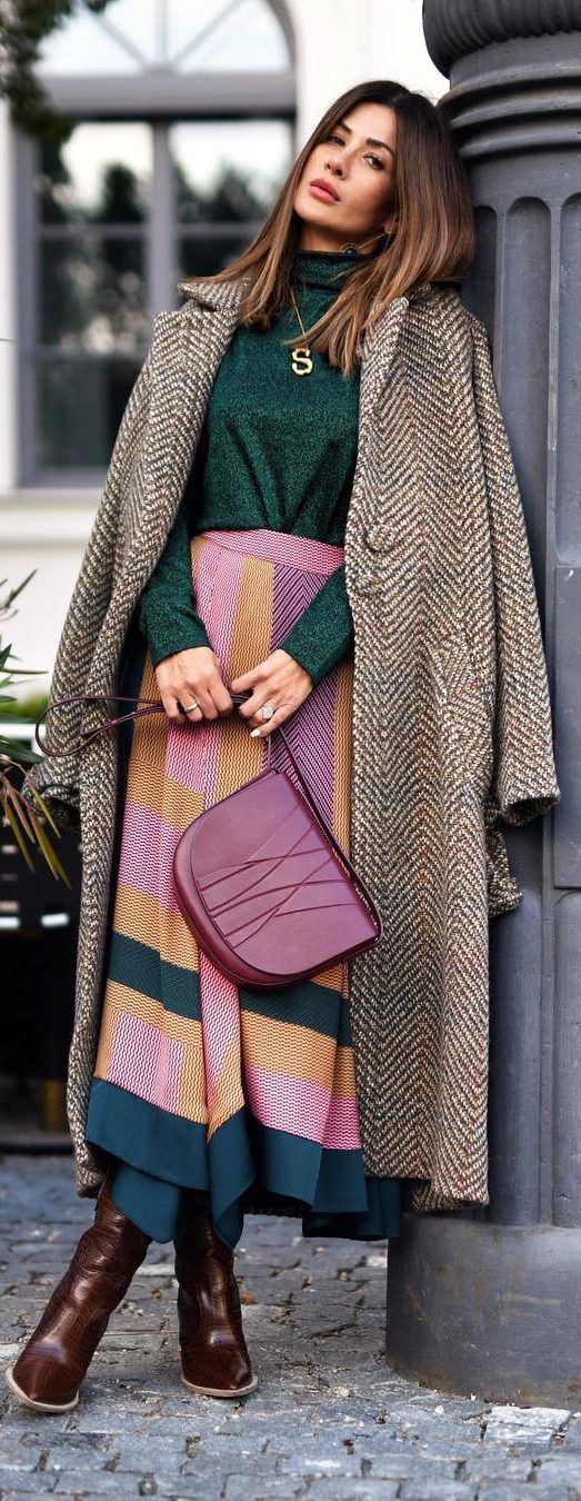 a stylish woman is posing against a stone pillar, showcasing her autumnal outfit. she wears a long herringbone coat over a green turtleneck sweater and a multicolored midi skirt.  the skirt has shades of pink, beige, and teal. she carries a burgundy handbag and is wearing brown ankle boots. her long brown hair is styled loosely.  the overall aesthetic is elegant and chic, representing a typical autumnal street style.