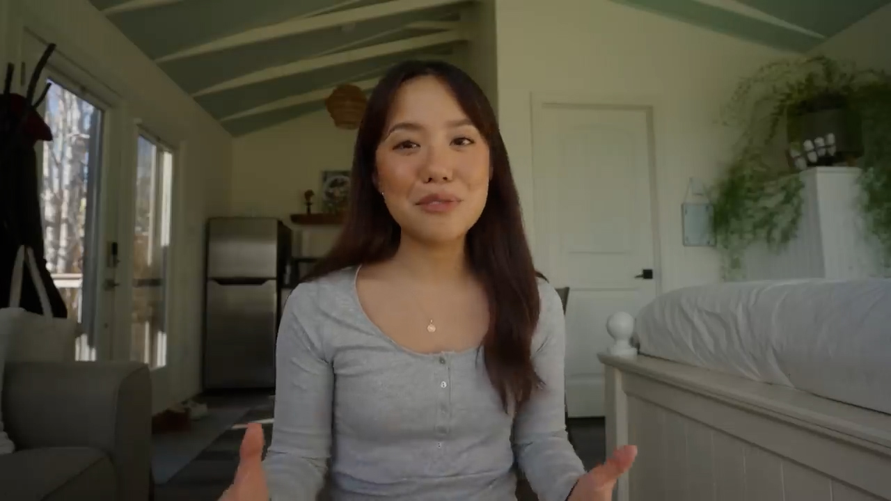 a young woman with long dark hair sits in a small, brightly lit room, possibly a tiny house or studio apartment. she is looking directly at the camera with a pleasant expression, as if speaking to an audience. the room is sparsely decorated in a minimalist style with light walls and simple furniture. a bed is visible in the background to her right, and a door and part of a kitchen area can be seen in the background behind her. the overall feel of the image is welcoming and casual.