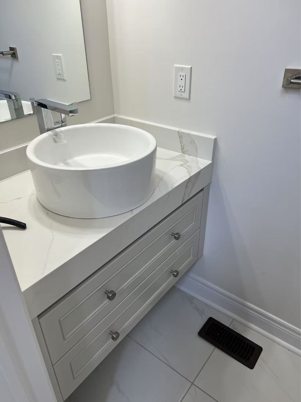 a bathroom features a round, white vessel sink atop a white vanity with a light gray marblepatterned countertop. the vanity has three drawers with brushed nickel knobs. a chrome faucet is mounted to the countertop behind the sink. an electrical outlet is on the wall above the countertop. a mirror is partially visible on the left side. a black vent is on the floor in front of the vanity. the floor is tiled with large, light gray tiles with a marble pattern. the walls are white.