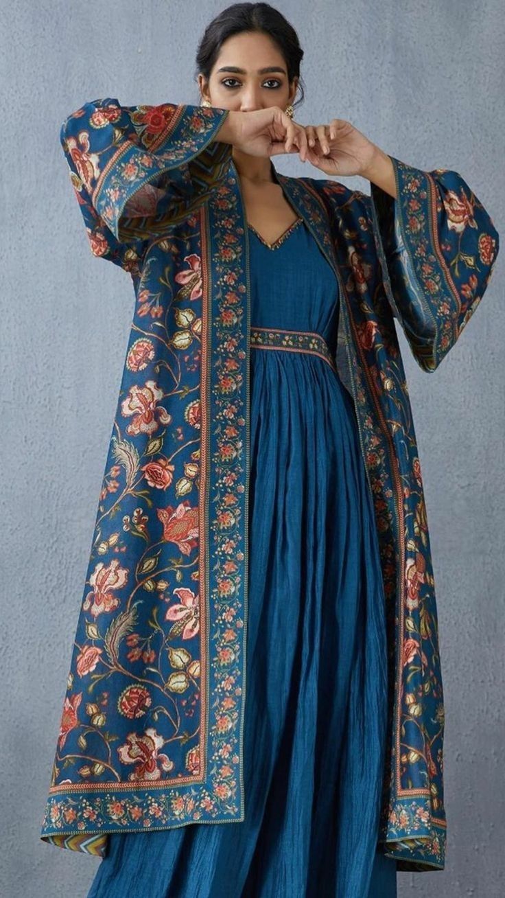 a woman is posing wearing a long teal blue dress and a matching floral printed coat. the coat has wide sleeves and a detailed border. the dress has a gathered waist and a simple neckline.  the overall style is elegant and bohemian, evoking an indian or ethnic fashion aesthetic. the background is a muted greyblue.