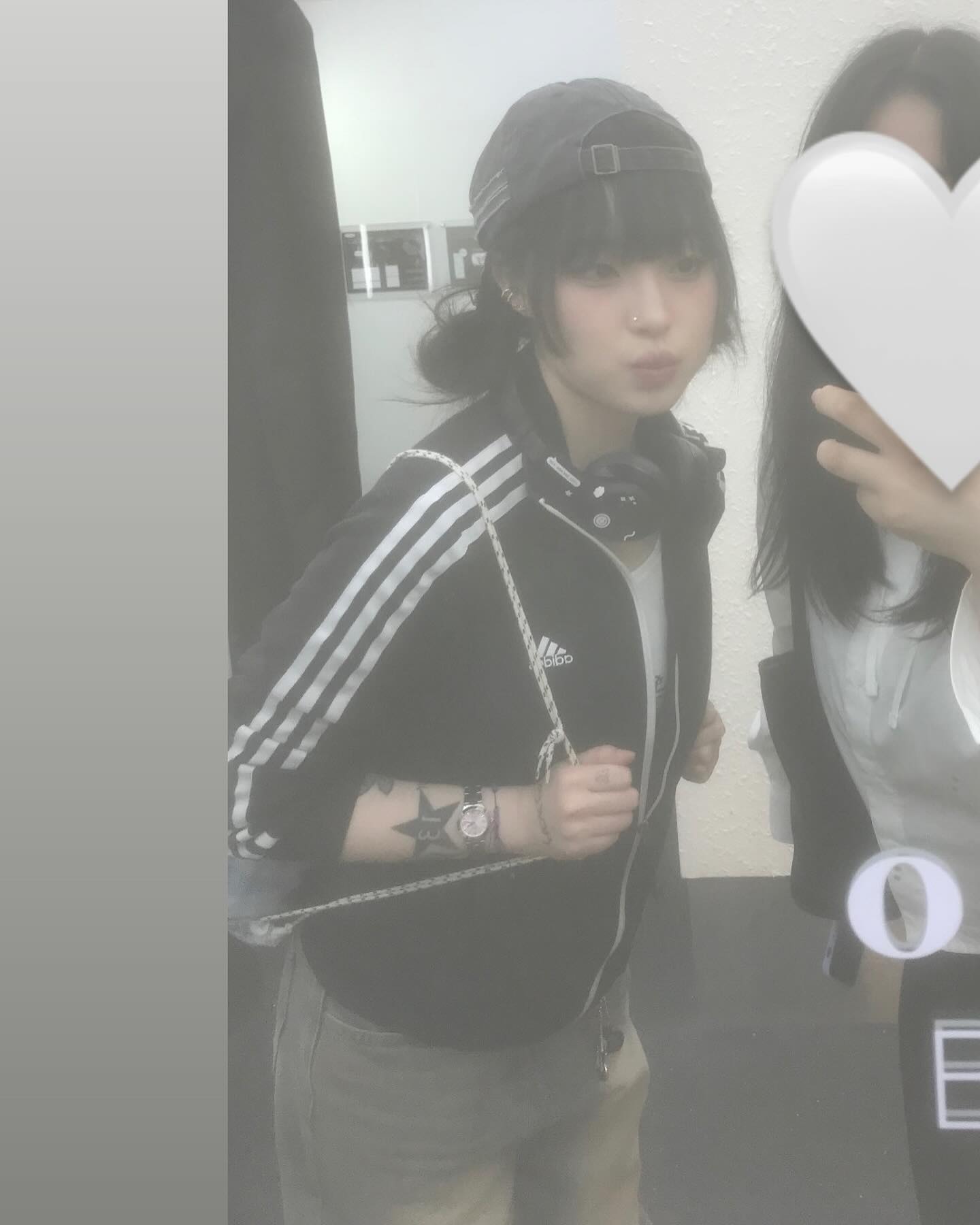 a young woman takes a mirror selfie. she's wearing a black adidas track jacket with white stripes on the sleeves, a black baseball cap, and jeans. she has a tattoo on her left arm and is wearing a watch. the photo has a slightly grainy and hazy effect.  a person holding a large white heart shape covers part of the right side of the image.