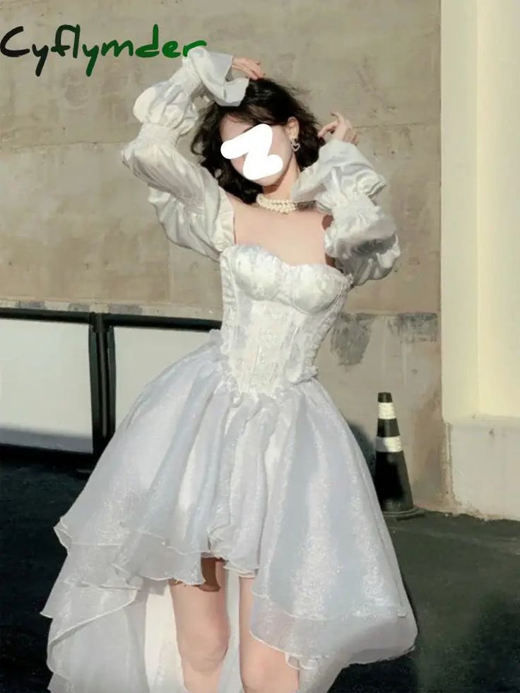 a woman is posing in a white dress with a corset bodice, puff sleeves, and a hilo hemline. she is also wearing a pearl necklace. the background is a building and a traffic cone.