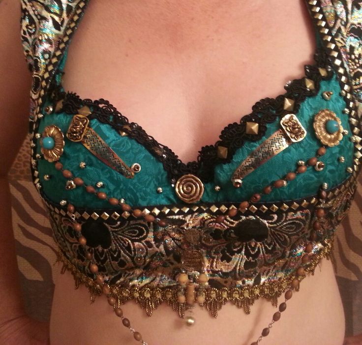 a closeup shot features a woman wearing an elaborate and colorful belly dance bra. the bra is teal and adorned with intricate details like black lace trim, gold studs, turquoise stones, and various metal embellishments. beads and coins dangle from the bottom of the bra, adding to its ornate and decorative appearance. the bra's design suggests a middle eastern or tribal aesthetic.