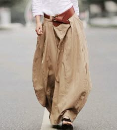 a woman is walking down the street. she is wearing a beige skirt, a white shirt, and a brown belt. her skirt is long and flowy. she is wearing a white shirt underneath. her belt is brown and is cinched at her waist. she is walking casually down the street and appears to be on her way somewhere.
