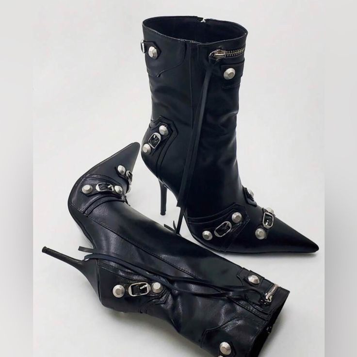 the image shows a pair of black leather ankle boots with a pointed toe and stiletto heels. the boots are adorned with silver embellishments such as studs and buckles. one boot is standing upright, while the other is lying on its side. the boots feature a zipper detail at the top and appear to be in a studio setting against a white backdrop.