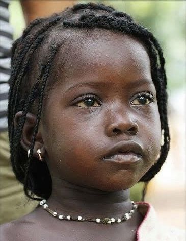a closeup portrait of a young african girl with dark skin and braided hair. she is wearing earrings and a necklace of white beads. she is looking slightly up and to the right.
