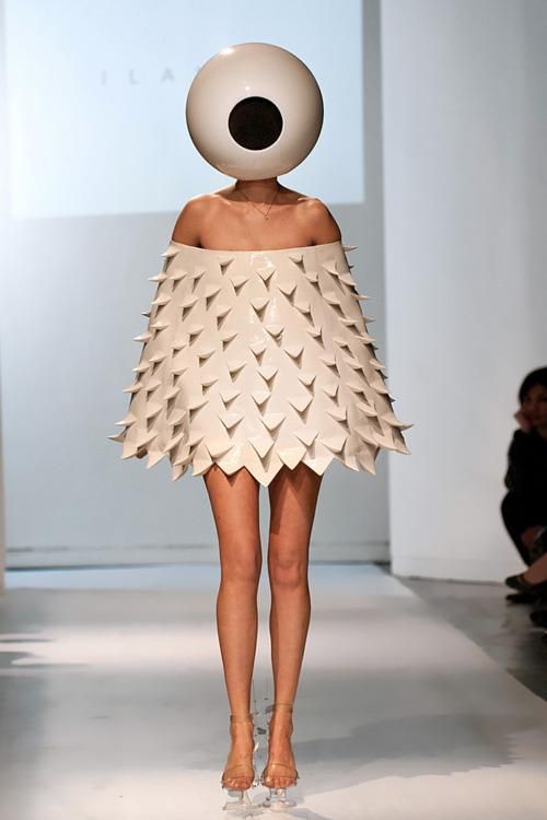 a model walks down a runway, wearing a unique and stylish outfit. her head is covered with a large, white, eyeshaped headpiece. she wears a short, white, dress with a unique, textured design that resembles a spiky sea urchin.  the model wears a pair of nude high heels. she looks at the camera with a confident and elegant pose.