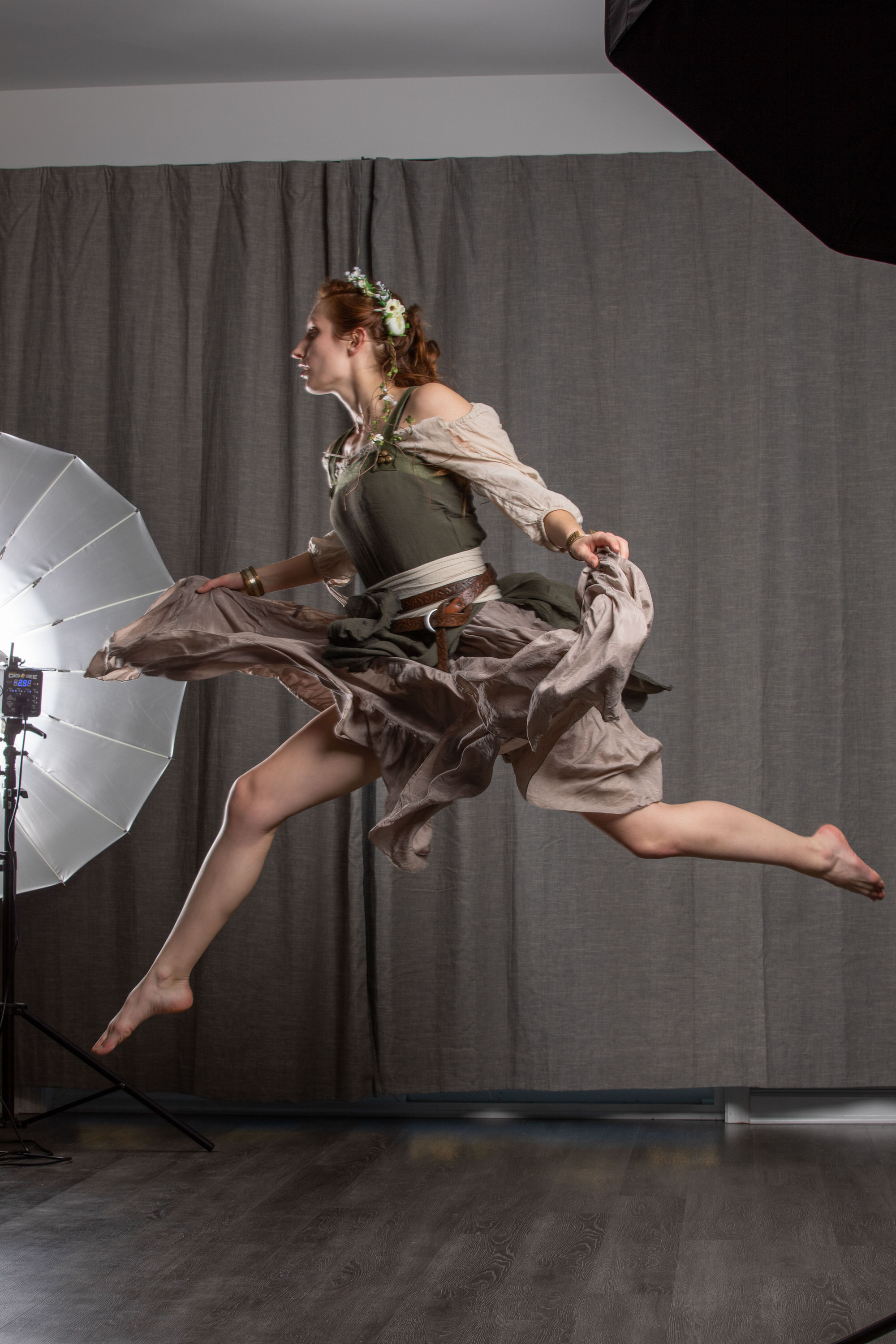 a young woman with reddish hair in a medievalinspired costume leaps through the air in a photography studio. she wears a flowing dress with a brown bodice and a lighter, sheer overskirt. her hair is adorned with a delicate floral crown.  two studio lights illuminate the scene, one with a softbox to the left and another, partially obscured, on the right. the backdrop is a simple grey curtain, and the floor is a dark wood laminate.