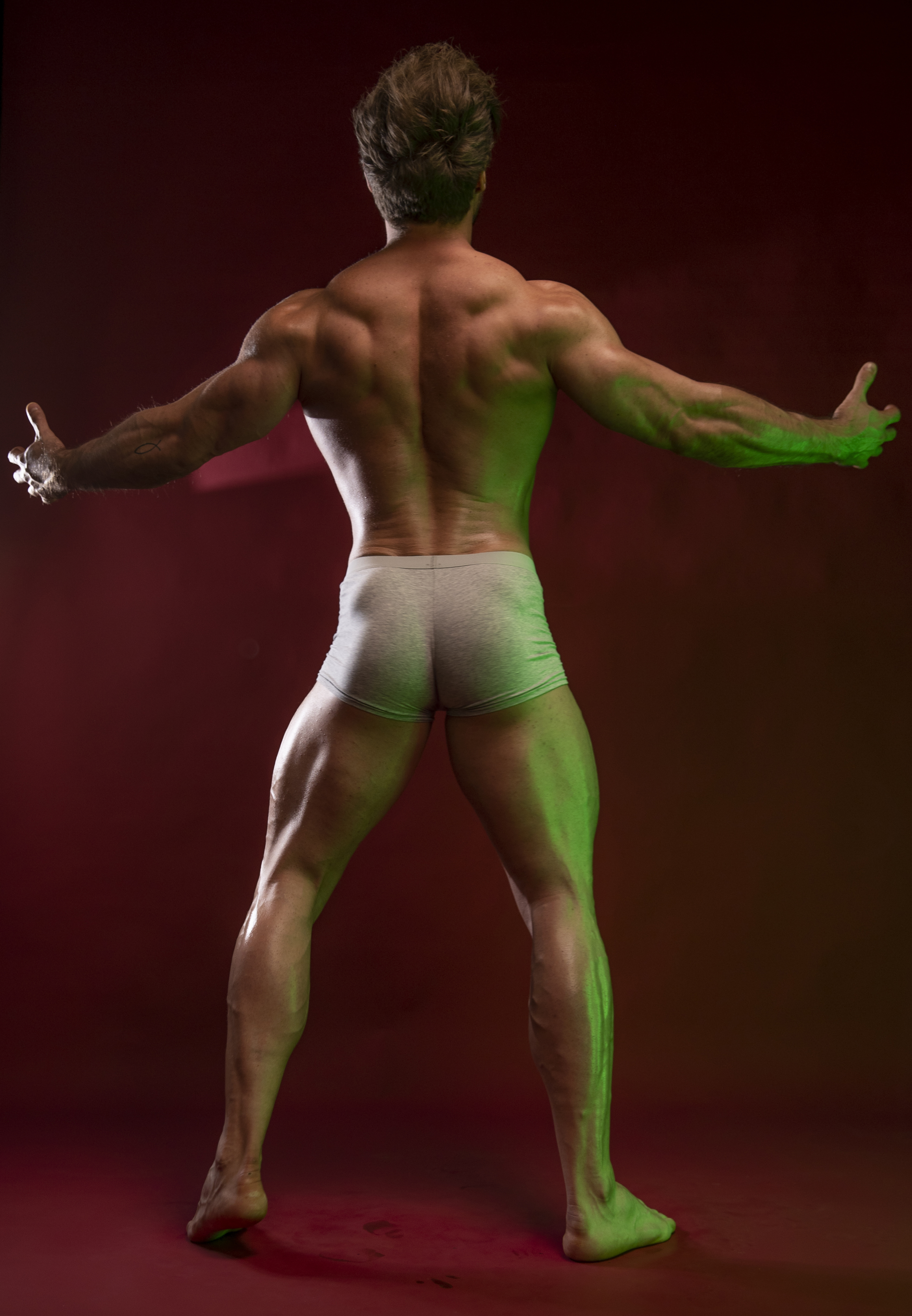 a muscular male bodybuilder stands with his back to the viewer. he is posing with his arms outstretched. he has welldefined muscles in his back, arms, and legs. he is wearing grey shorts. the background is a dark red color, and green light is shining on his body.