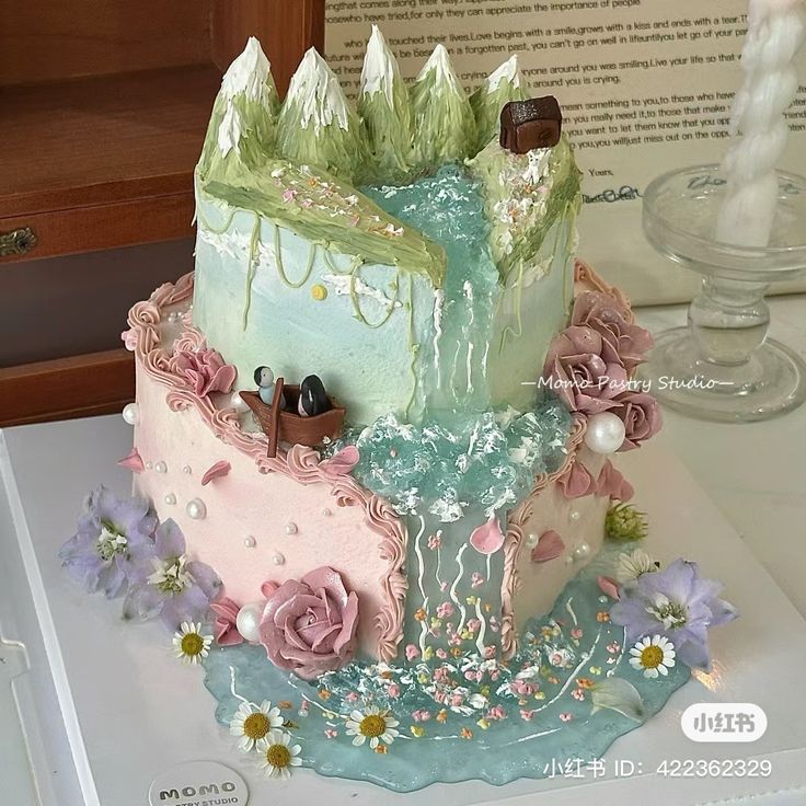 the image shows a stunning twotiered cake created by momo pastry studio. the cake is designed with a whimsical landscape theme, featuring pastelcolored buttercream frosting, delicate floral decorations, pearl accents, and miniature figurines on a small boat. the top tier depicts a mountain scene with a waterfall cascading down to the bottom tier which includes a lake. the entire cake is exceptionally detailed, with a beautiful color palette and textures giving it a romantic and dreamy aesthetic. the cake sits on a white surface, with the studio's logo visible at the bottom.  the image also includes a small portion of text visible in the background and a small red logo in the bottom right corner with a social media id.