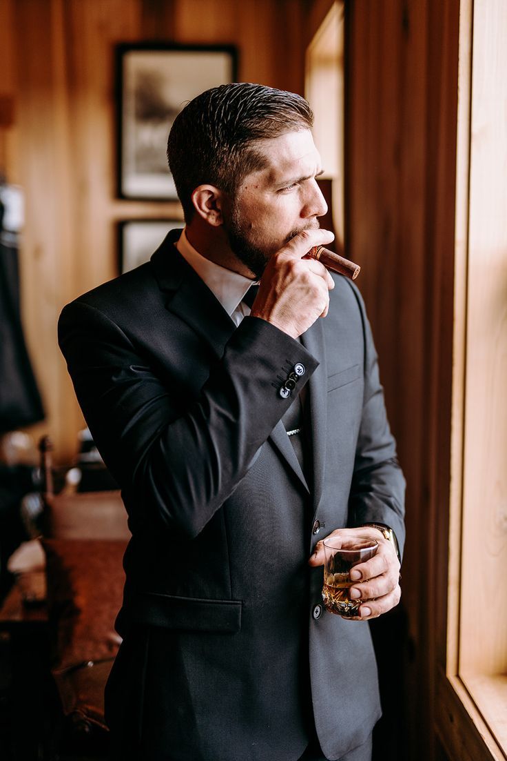 a man in a black suit stands by a window in a wooden room. he is smoking a cigar and holding a glass of whiskey. he looks serious and thoughtful.