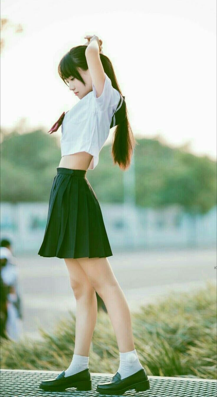 a young woman in a school uniform is standing in an outdoor setting. she is wearing a shortsleeved white shirt, a pleated black skirt, white socks, and black loafers. her hair is long and dark. she has her hands up near her head.