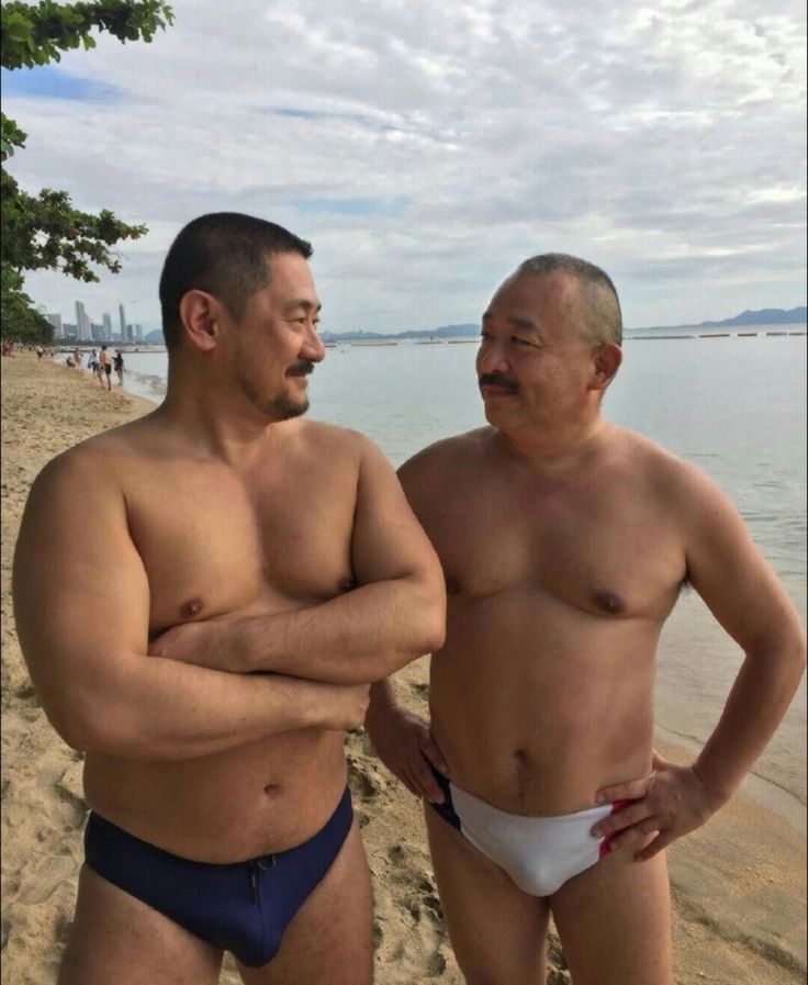 two men are standing on a sandy beach with the ocean behind them. they are both smiling and looking at each other. they are both wearing swim trunks. the man on the left is wearing dark blue trunks and the man on the right is wearing white trunks with a red stripe. they appear to be friends enjoying a day at the beach.