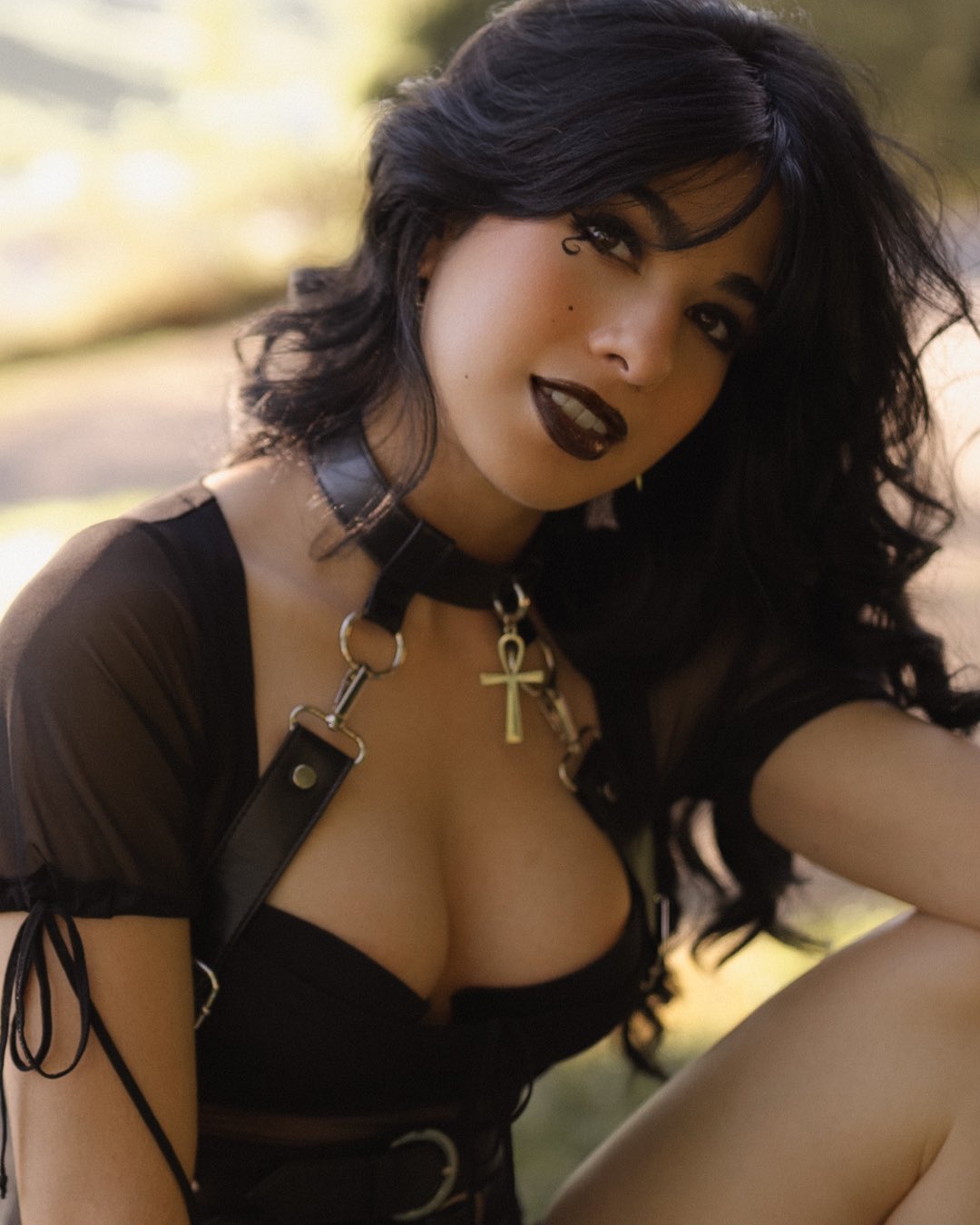 a portrait of a woman with long black hair, dark lipstick, and distinctive makeup, including a beauty mark near her eye. she is wearing a black collar and a harness with an ankh pendant. she has on a low cut top with sheer sleeves. the setting appears to be outdoors, likely in a park or wooded area. she is sitting down, with a portion of her legs visible. the overall style leans towards a goth aesthetic.