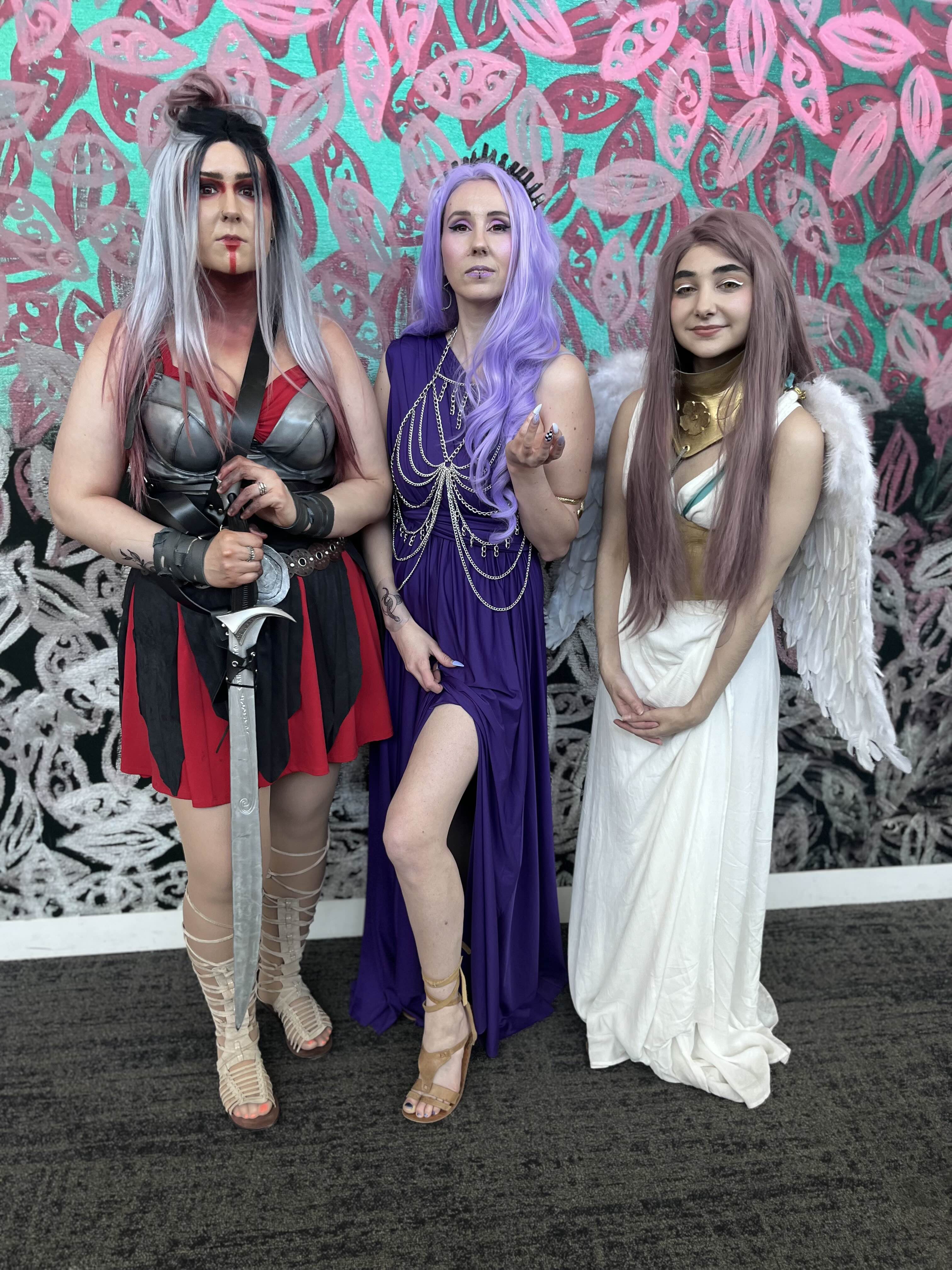 three women in elaborate fantasy cosplay costumes are posing against a vibrant wall with floralpatterned artwork.  one woman is dressed as a warrior with a sword, wearing a red and grey outfit and gladiator sandals.  another is in a long purple dress with a jeweled neckline. the third is costumed as an angel, wearing a flowing white gown and feathered wings. they are smiling and appear to be at a convention or similar event. the overall aesthetic is vibrant and whimsical.