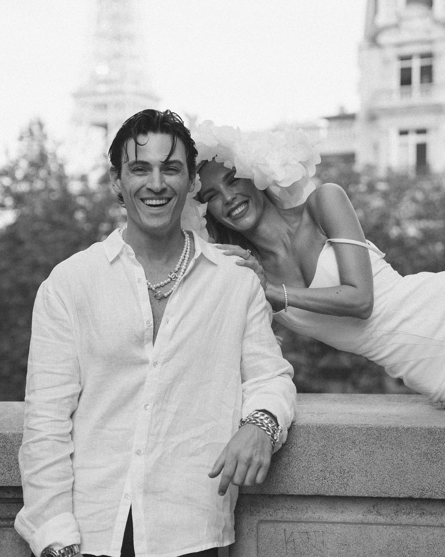 a black and white portrait of a couple smiling and looking at the camera. the man is wearing a white button down shirt and the woman is wearing a white dress with a white flower on her head. the couple is standing on a bridge with the eiffel tower in the background. they look very happy and in love.