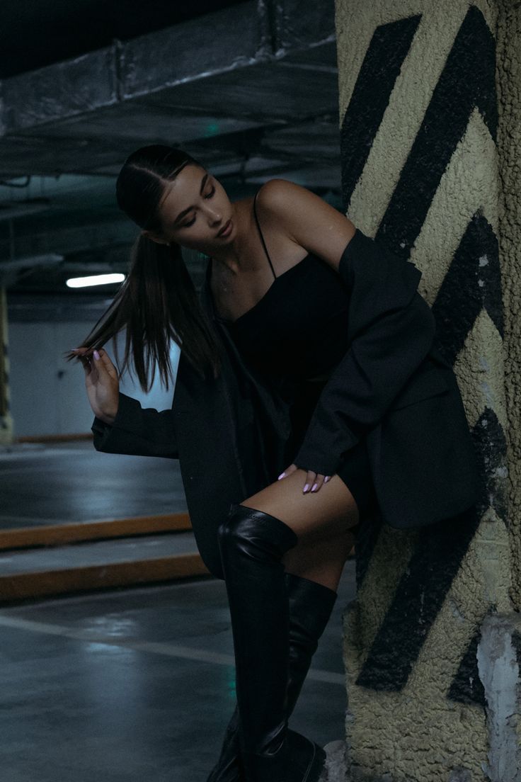 a young woman with long brown hair styled in a ponytail is posing in a parking garage. she is wearing a black dress, black blazer, and thighhigh black leather boots. she leans against a concrete pillar with black and yellow stripes. the overall atmosphere is dark and moody. the image has a fashion and editorial feel.