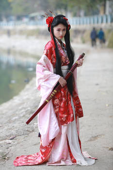 a young woman in a beautiful red and pink kimono stands outdoors holding a katana. she is posed gracefully and her clothing is detailed and elegant. the background is slightly blurred, but shows a parklike setting near water.