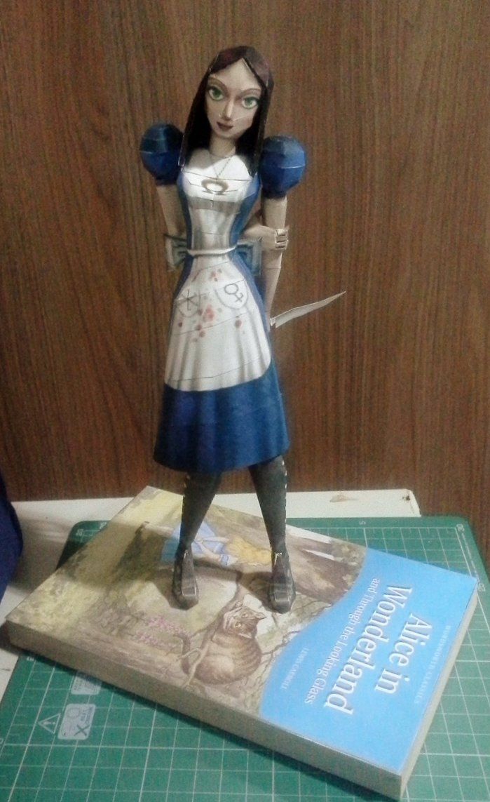 meticulously crafted papercraft figurine of alice from alice in wonderland stands on a copy of the book. the figurine is highly detailed, showcasing alice's iconic dress and boots. she holds a knife, adding a dramatic touch to the pose. the background is a simple wooden surface, and the image is welllit, highlighting the intricate details of the papercraft. the book adds a context to the scene.