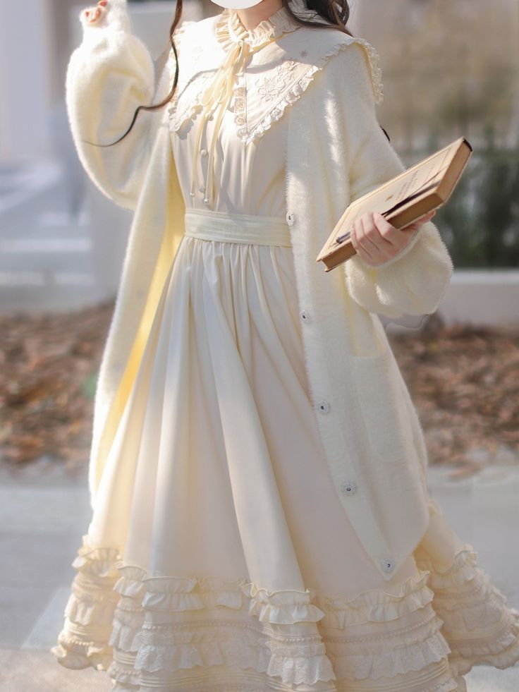 a person is standing outside wearing an ivory dress with lace detailing and ruffles at the hem. they are also wearing a fuzzy white cardigan and holding a book in their left hand. the photo is taken from the waist down, with the person's face not visible. the lighting is natural and soft, giving the image a warm and inviting feel.