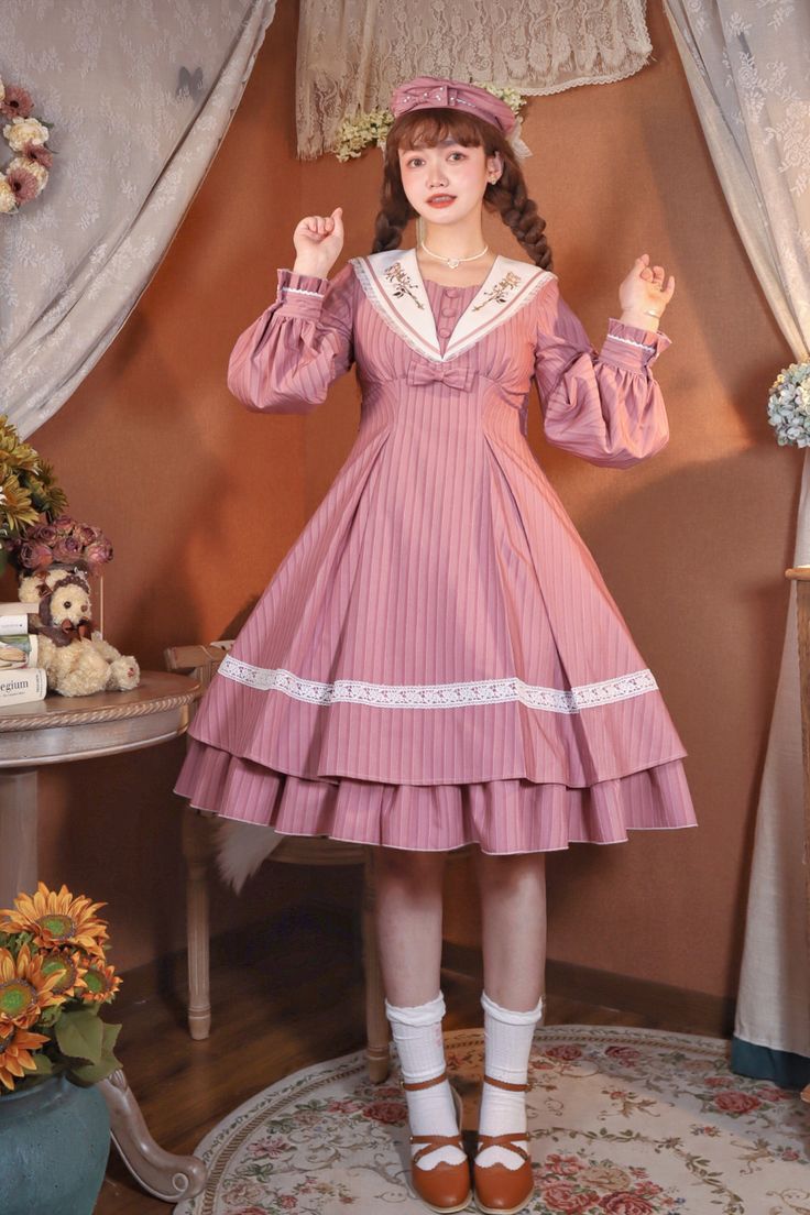 a woman is posing in a pink and white lolita fashion dress. she is wearing a matching hat and her hair is in braids. she is standing in front of a curtain and there is a table with flowers and a teddy bear on it to her left. the overall color palette is soft and feminine.
