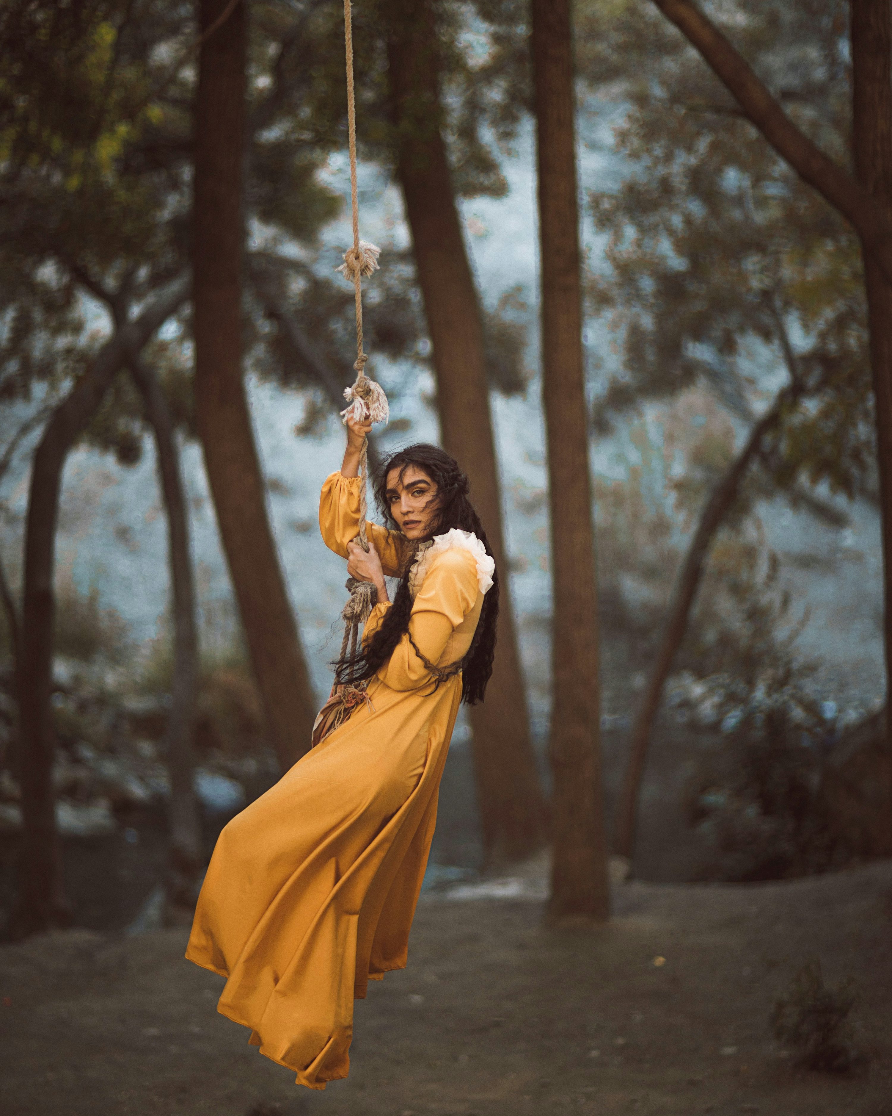 a young woman with long dark hair is captured midswing on a rope swing in a wooded area. she wears a flowing, long yellow dress that billows around her as she swings. the setting appears to be a forest with tall, slender trees, suggesting an autumnal or fall atmosphere. the overall mood of the image is dreamy, whimsical, and slightly romantic. the lighting is soft and diffused, contributing to the ethereal quality of the photograph. the focus is sharp on the woman, while the background is slightly blurred, drawing the viewer's attention to her. the image has a cinematic and artistic feel.