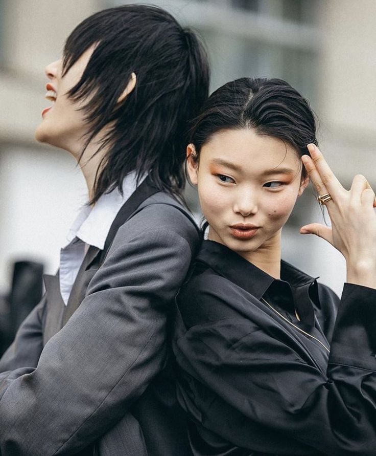 two stylish models are looking at the camera while standing back to back. the model in the back is wearing a black suit jacket and white collared shirt. the model in the front is wearing a black shirt with a collar. they both have trendy haircuts. this photo was taken outdoors.