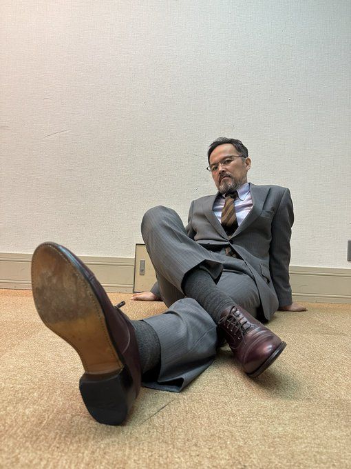 a middleaged man in a welltailored grey suit and polished brown dress shoes sits confidently on the floor, leaning back on his hands. he makes direct eye contact with the camera, projecting an air of ease and selfassurance. the simple background and low camera angle emphasize his presence.