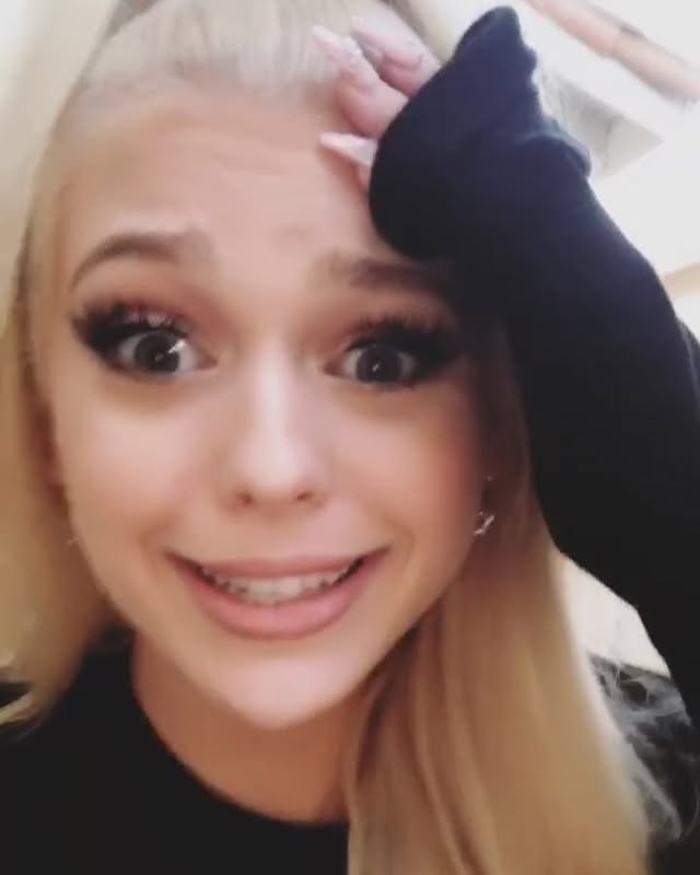 a closeup shot of a young woman with blonde hair and fair skin. she has a surprised or worried expression on her face, with wide eyes and a slight smile that reveals braces on her teeth. she's wearing makeup, including eyeliner and mascara. her hand is raised to her forehead. the image has a slightly grainy or soft focus quality.