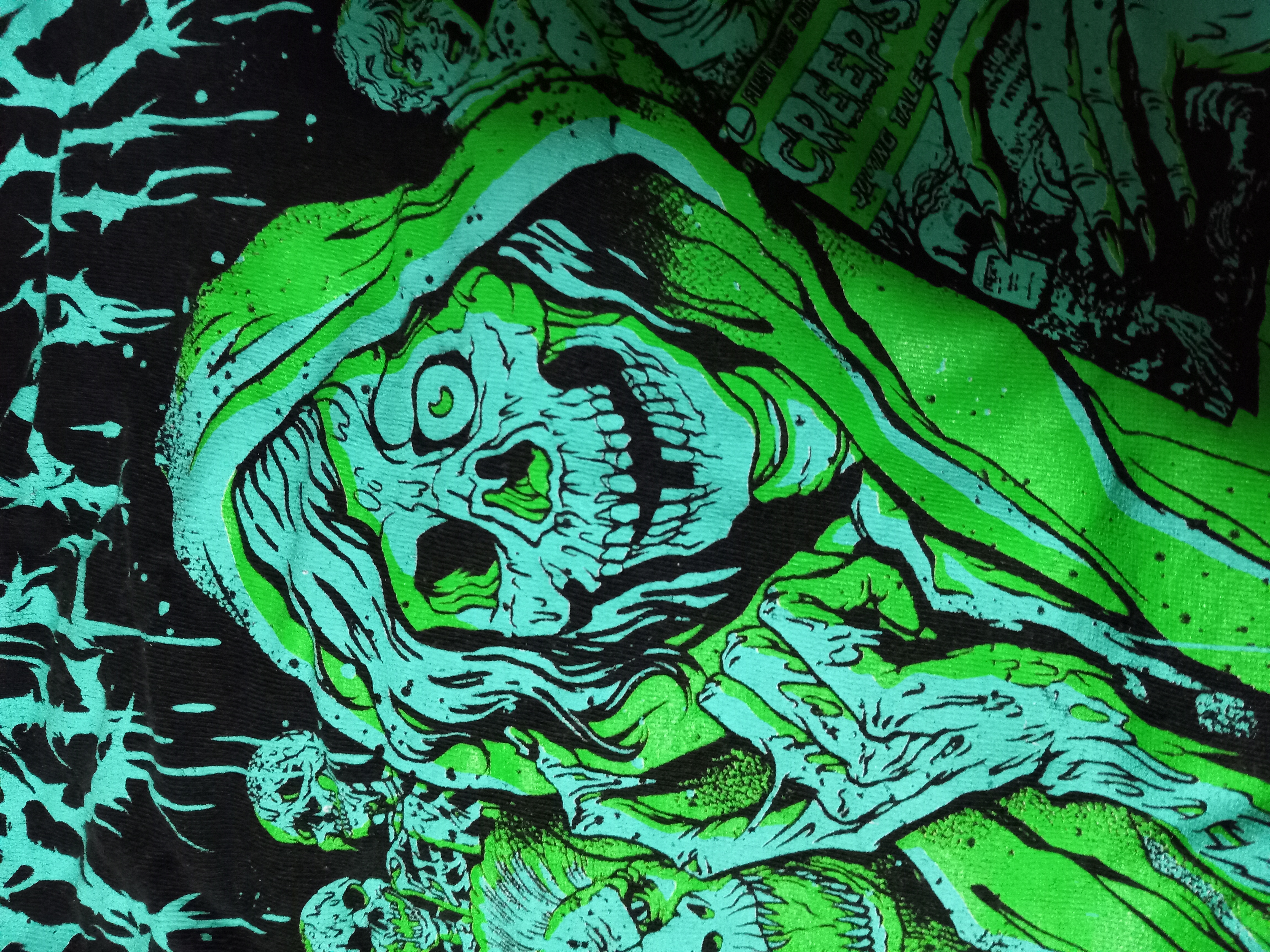 loseup shot of a black tshirt with a vibrant green and black graphic design. the design features a central, hooded figure resembling a skeleton or zombie with decaying flesh.  surrounding the main figure are other smaller skulls and skeletal figures, creating a chaotic and macabre scene. the style is reminiscent of horror movie posters, comic books, or heavy metal band merchandise. the text "creeps" is partially visible on a comicbook style object held by the main figure. the overall aesthetic is dark, gritty, and visually striking.