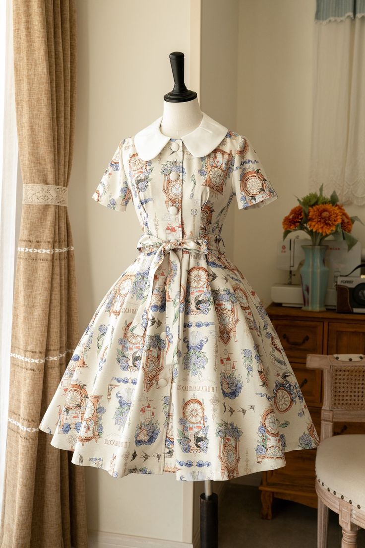 this is an image of a creamcolored dress with a floral and clock print. the dress is displayed on a mannequin in a welllit room. the dress has short sleeves, a collar, and an aline skirt. the background includes a curtain, a vase of flowers, a chair, and a mirror.