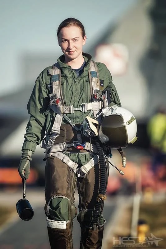a female pilot in a flight suit and helmet walks towards the camera on the flight line. she is carrying her helmet under her arm and has a microphone on her chest.