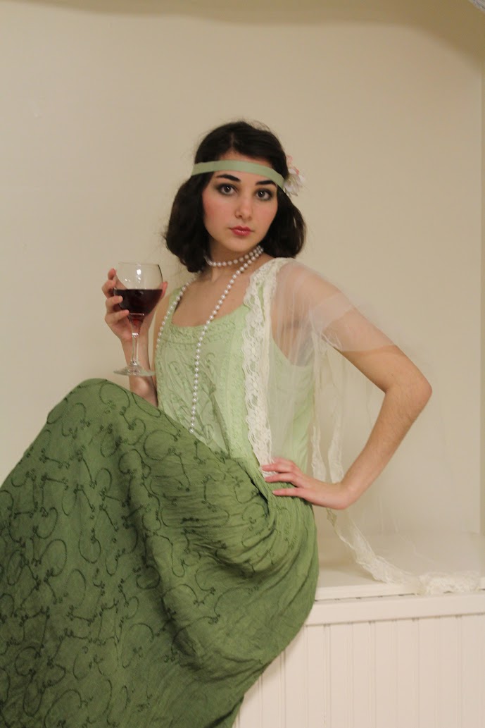 a young woman, dressed in a vintage 1920s flapper dress, sits on a white wooden surface. the dress is a pale green, with lace detailing on the bodice and an embroidered green skirt. she is accessorized with a pale green headband, a pearl necklace, and holds a glass of red wine. the background is a plain, offwhite wall. the woman's expression is subtle and somewhat reserved, creating an air of elegance.  the overall image evokes a sense of vintage fashion and the flapper era.