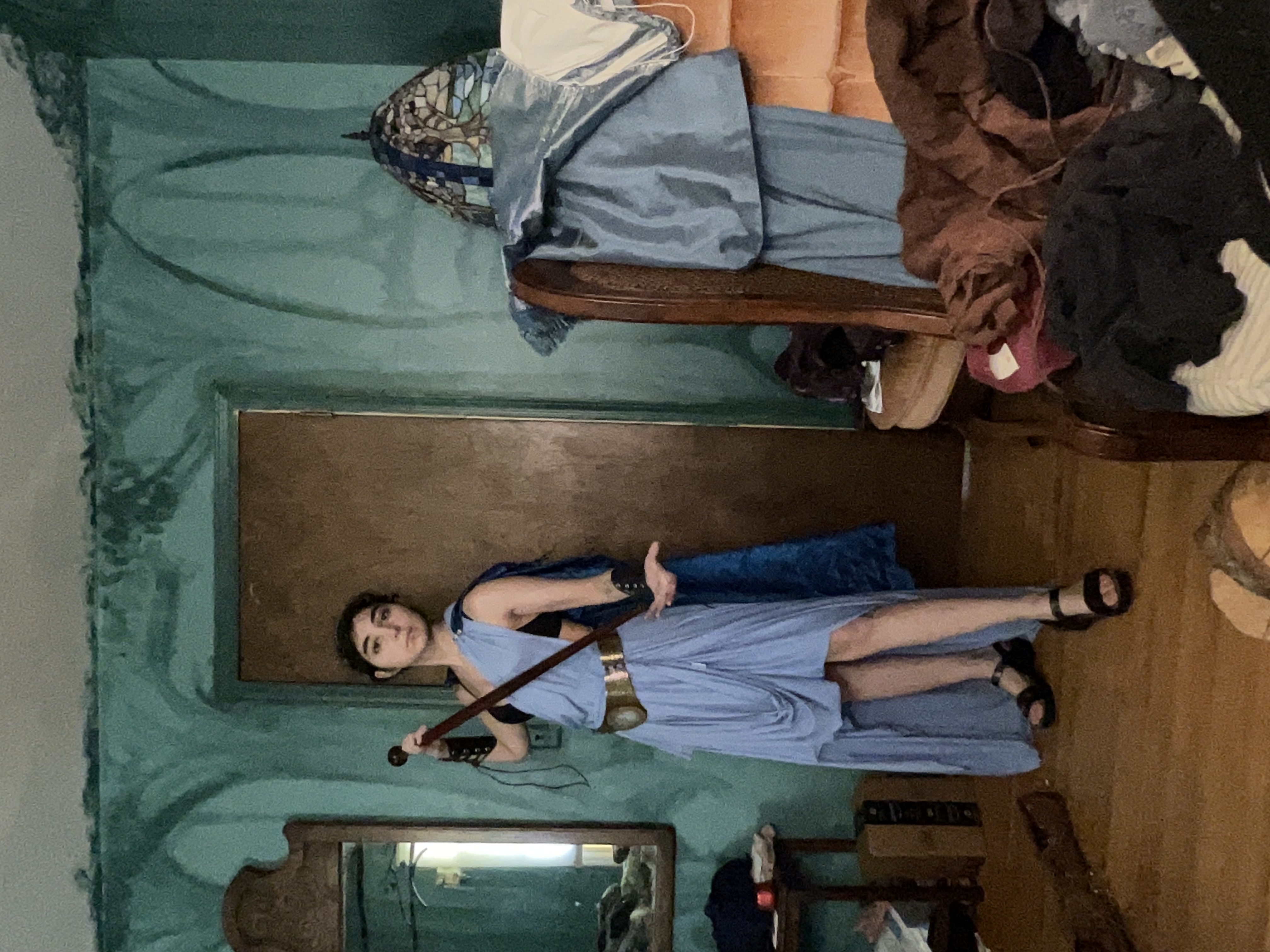 a woman in a blue highlow fantasy warrior cosplay stands in a cluttered room with teal painted walls. she holds a staff and is wearing sandals. the room features a vintage chair, a mirror, and various items scattered around. the overall aesthetic is retro or vintage, possibly reminiscent of the 1970s or 1980s. the woman's costume appears to be handmade, and she is posing for a picture.  the background is somewhat messy and untidy.