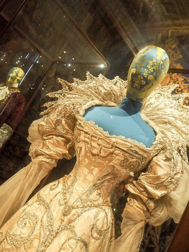 a close up of an elaborate gold dress with lace and embroidery on display in a museum case. the dress is worn by a mannequin and appears to be a historical piece of clothing. the dress has a large, ornate collar and long sleeves and is decorated with gold beads, sequins and lace. the dress is likely a royal gown from a past era.