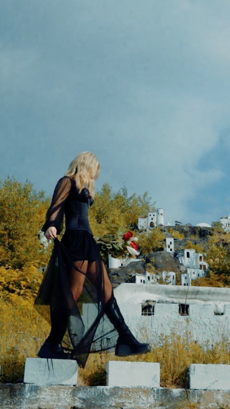 a woman in a black dress and platform boots walks down steps in front of a village. she is holding flowers. the sky is blue.