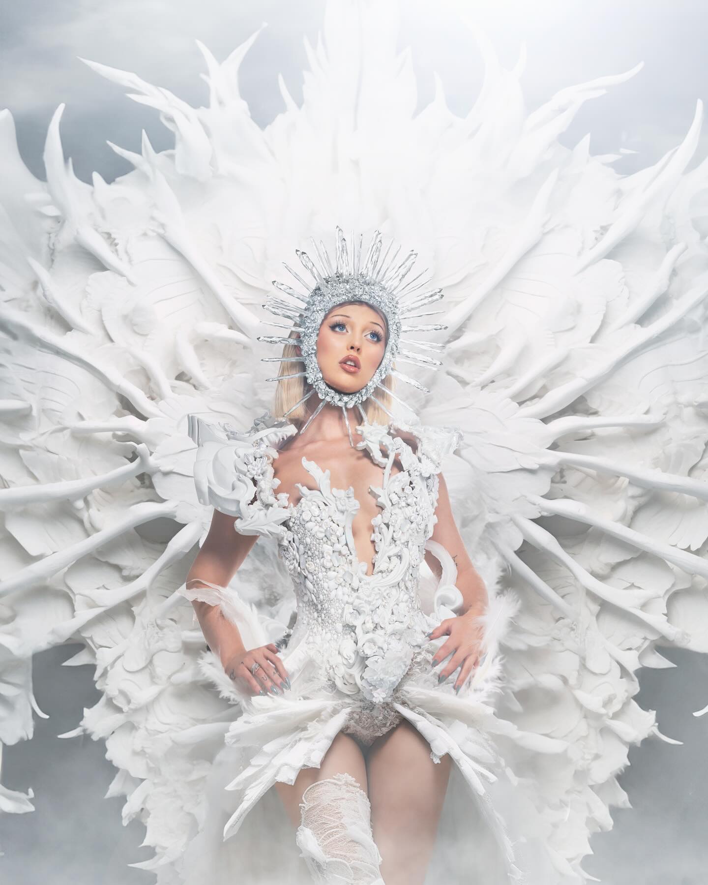 a woman stands in a full white costume resembling an angel. she is wearing a fitted, lowcut corset with elaborate feather and lace detailing, a large feathered winglike structure extending from her shoulders, and a silver crownlike headdress. the lighting is soft, giving the impression of ethereal beauty. the woman has blonde hair, elegant makeup, and is captured in a portraitstyle pose.