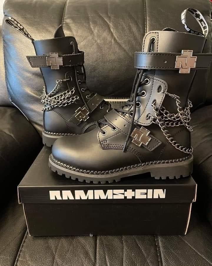 a pair of black leather rammstein boots are displayed on a black box. the boots are detailed with silver metal rammstein logos and chains.  the box is also branded with the rammstein logo. the boots are laceup style combat boots, appearing to be a collaboration or limited edition item.  they rest on a dark leather couch or chair.