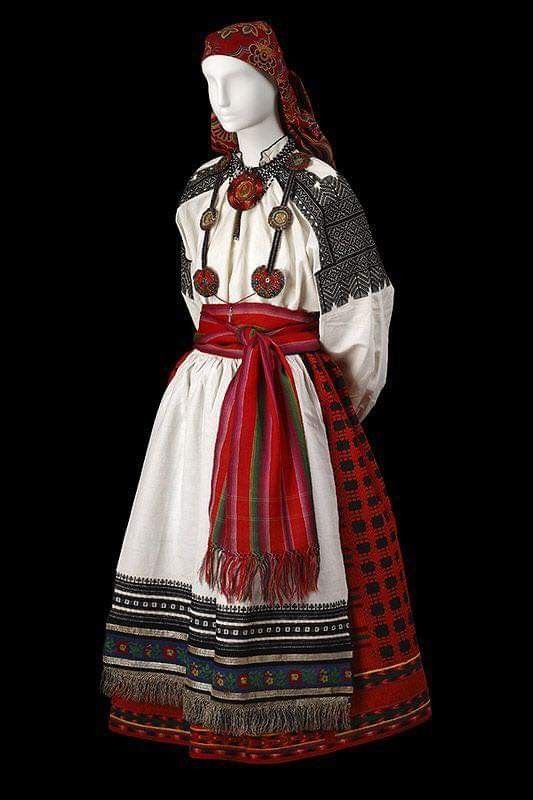 a mannequin wearing traditional eastern european clothing. the outfit includes a red and patterned headdress, a white dress with embroidered sleeves and hem, a red sash, and decorative jewelry. the clothing features intricate patterns and fringe details. the background is black.