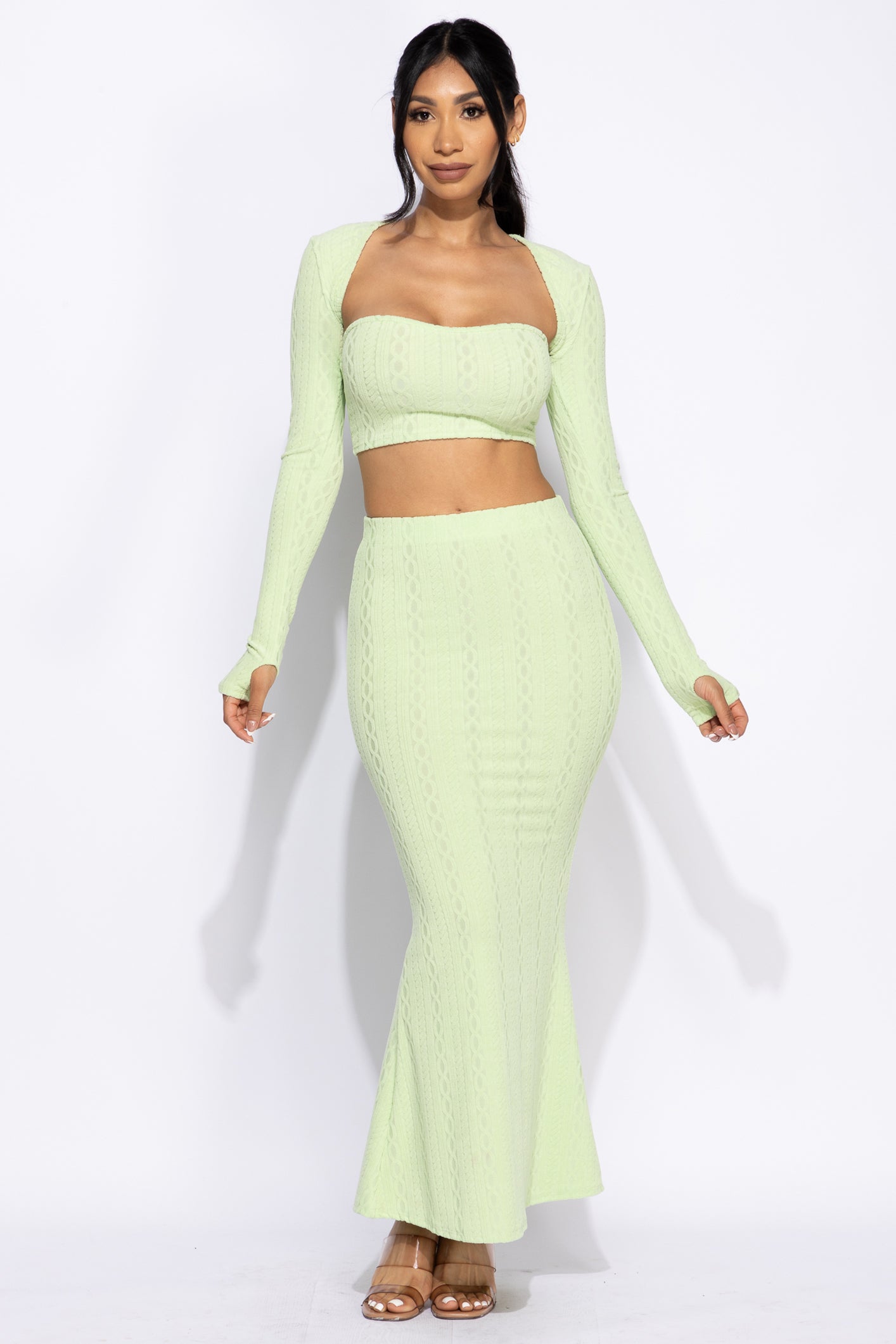 a full shot of a female model posing in a stylish lime green twopiece outfit. the outfit consists of a longsleeve knit crop top and a fitted maxi skirt with a mermaidstyle hem. the fabric has a subtle textured pattern. the model is wearing neutraltoned sandals. the background is plain white.