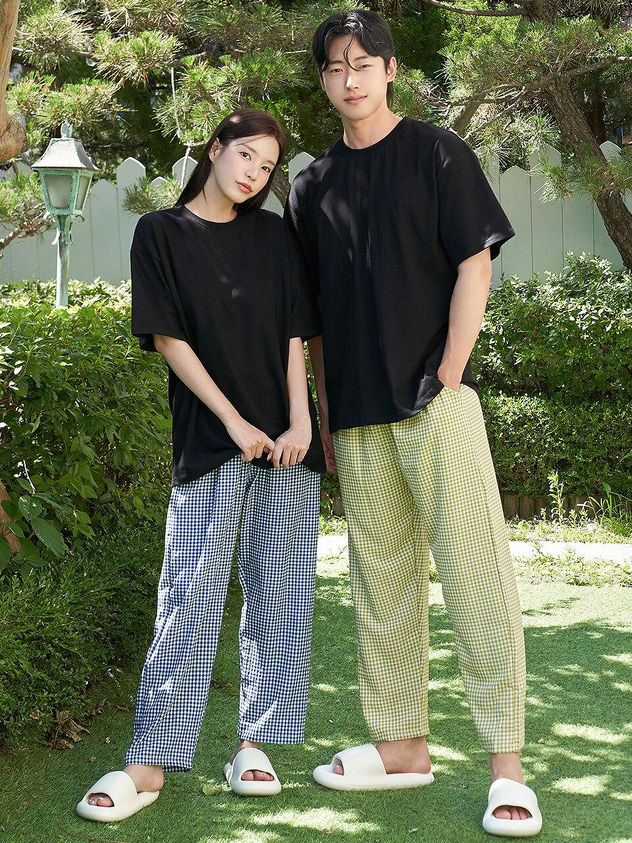 a couple is standing sidebyside outdoors. the woman is wearing a black tshirt and blue checkered pants, while the man is wearing a black tshirt and yellow checkered pants. both are wearing slides. they are standing on a grassy lawn with trees and bushes in the background.