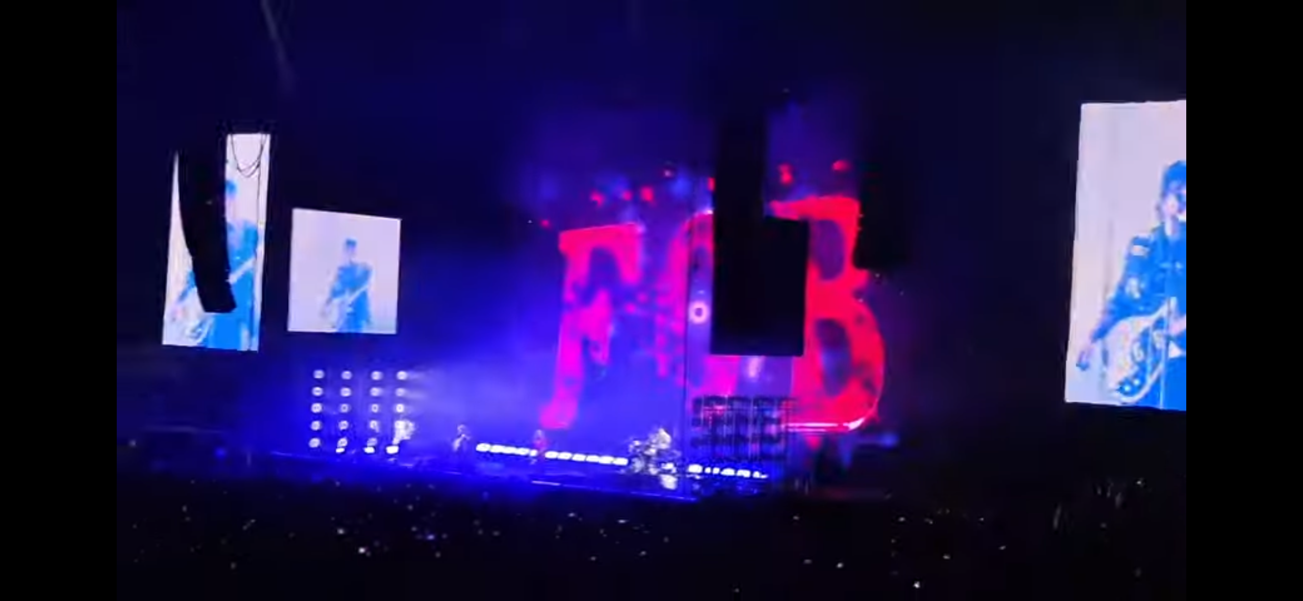 a concert stage with a large crowd in the background. the stage is lit up with bright lights and there are several large screens behind the band showing images of the band playing.  the band is playing music and the crowd is cheering.  the band's logo can be seen on the screen behind the band.