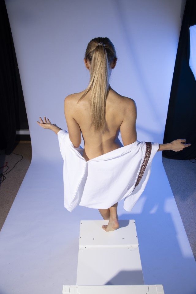 a woman is standing with her back to the camera. she has blonde hair in a ponytail. she is nude and is holding a white towel with a decorative border around her body. she is standing on a white platform in a studio with white and blue lighting.