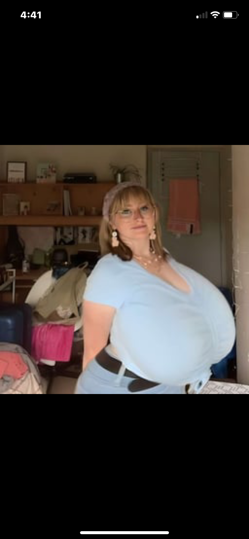 the image shows a smiling, plussize woman with large breasts wearing a light blue vneck shirt and jeans. she's posing confidently in what appears to be a dorm room or bedroom setting. she has on glasses and a headband.  the overall impression is one of body positivity and selfacceptance.