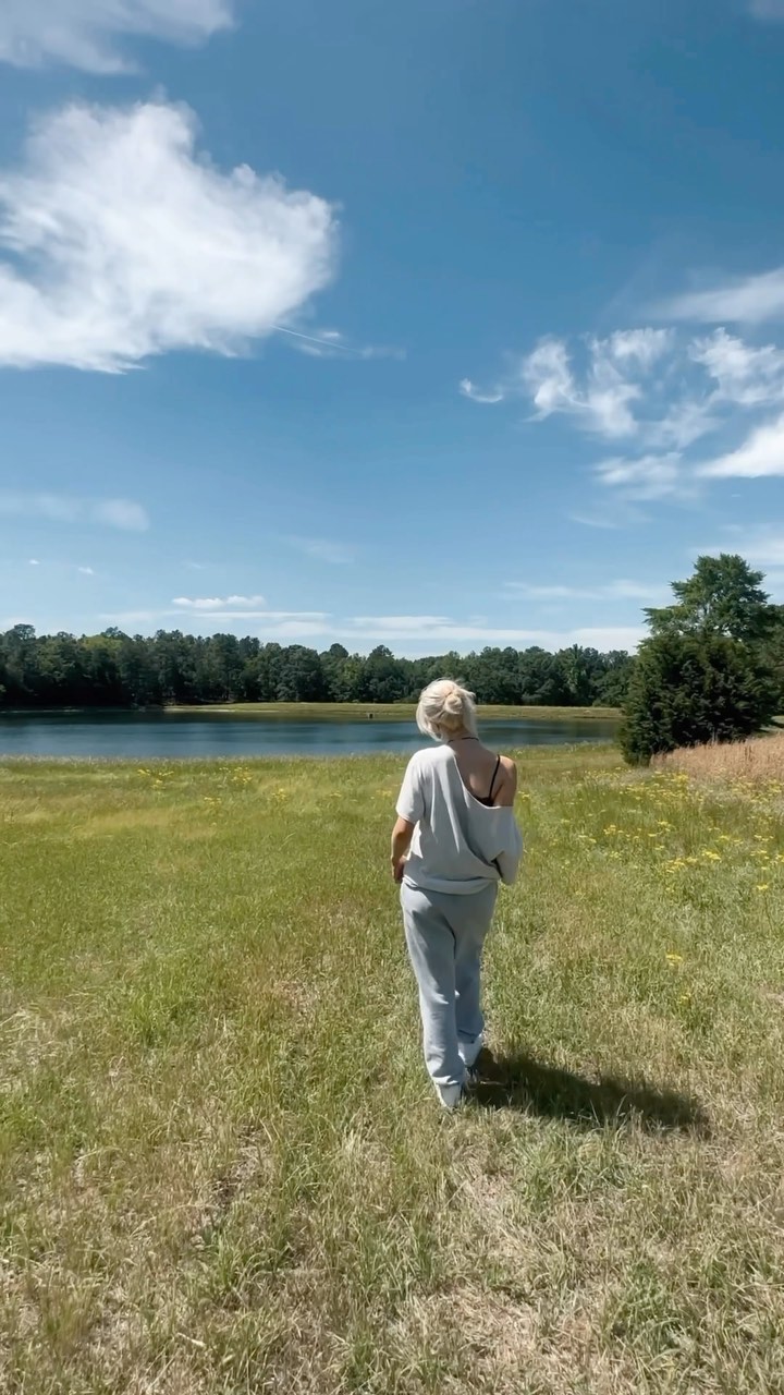 a woman walks through a grassy field towards a lake on a sunny day. the sky is blue with fluffy white clouds. trees line the far side of the lake. the woman is wearing casual clothing and has blonde hair. she is seen from the back, casting a shadow on the grass.