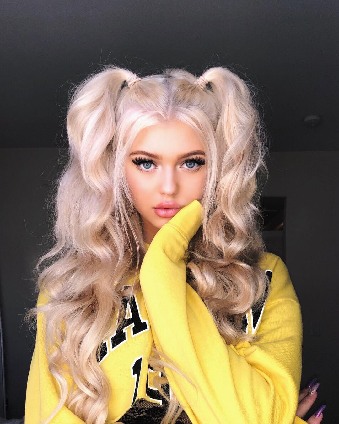 a woman with blonde hair styled in pigtails, wearing a yellow sweater, is posing indoors. she has blue eyes and wears makeup.